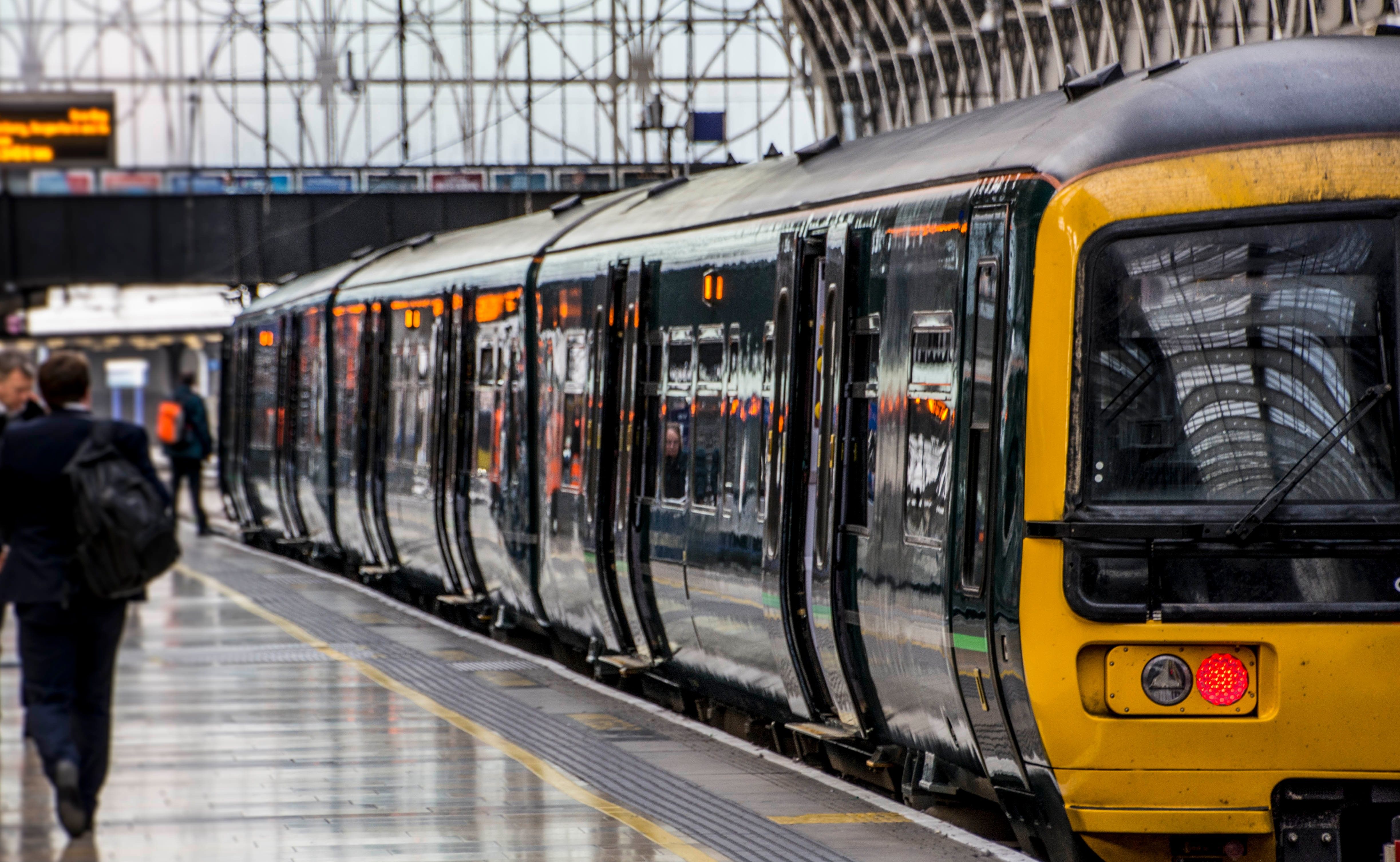 A modern passenger train with a yellow front is stopped at a busy platform inside a large covered railway station, with people walking alongside the train